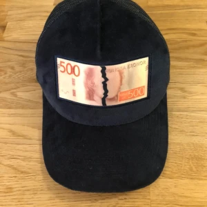 Marinblå keps med 500-lapp patch från Bruce Lee x All Cap Co - Cool marinblå keps med mesh på sidorna och en unik patch framtill som föreställer en trasig svensk 500-kronorssedel. Justerbar baktill och broderad logga på sidan. Perfekt statement-piece för dig som gillar streetwear. Perfekt till sommaren