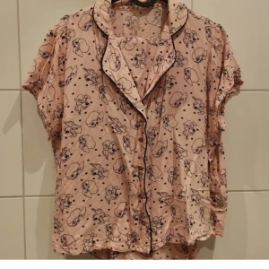 Rosa pyjamasskjorta med hundmönster - Söt rosa pyjamasskjorta med svarta hundmotiv och små hjärtan. Skjortan har korta ärmar, svarta kantband och knappar framtill. Perfekt för mysiga hemmakvällar.