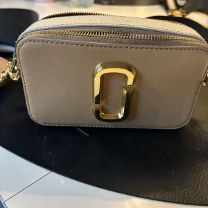 Beige axelväska från Marc Jacobs med gulddetaljer - Säljer en beige axelväska från Marc Jacobs med guldfärgad logga framtill och dubbla dragkedjor. Väskan har svart bakficka och justerbar axelrem. Perfekt för att bära det viktigaste med stil. det finns ”spår” av läppglans på insidan av väskan. Orginalpåsen finns kvar 