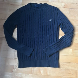 Mörkblå kabelstickad tröja från GANT - Säljer en klassisk mörkblå kabelstickad tröja från GANT med rund halsringning och broderad logga på bröstet. Tröjan har långa ärmar och ribbade muddar vid ärmslut och nederkant. Perfekt för lager-på-lager under kyliga dagar.