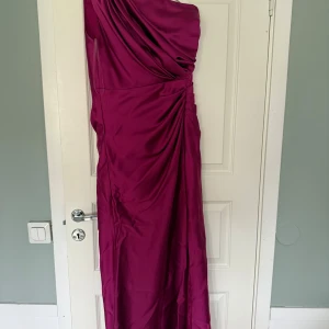 Mörkrosa långklänning med oneshoulder och slits - Säljer en elegant mörkrosa långklänning med oneshoulder och draperad överdel. Klänningen har en snygg slits framtill och medföljer ett bälte med glittrigt spänne. Perfekt för bal eller festliga tillfällen. Används endast en gång så inget fel på den!