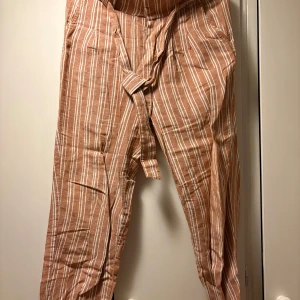 Randiga linnebyxor med knyt från Gina Tricot - Säljer ett par snygga, brunbeige linnebyxor från Gina Tricot i storlek 36. Byxorna har vita ränder, knytband i midjan och avslappnad passform. Perfekta för varmare dagar och ger en chill vibe till outfiten.