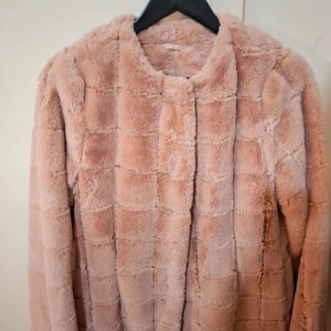 Ljus rosa fuskpälsjacka från Visual Clothing Project - Mysig ljus rosa pälsjacka med rutstruktur från Visual Clothing Project. Jackan har dold knäppning framtill och är helfodrad. Perfekt för kyliga dagar när du vill känna dig extra stylish.