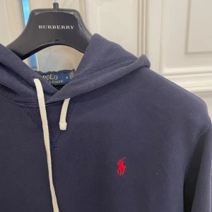 Mörkblå hoodie från Polo Ralph Lauren - Säljer en klassisk mörkblå hoodie från Polo Ralph Lauren med röd broderad logga på bröstet. Tröjan har huva med vit dragsko och en stor magficka framtill. Perfekt för en avslappnad och stilren look.