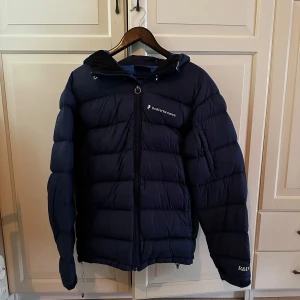 Jacka - Säljer en mörkblå dunjacka från Peak Performance med huva och dragkedja framtill. Jackan har en pufferstil och logga på bröstet samt ärmarna. Perfekt för kyliga dagar och riktigt skön att bära.