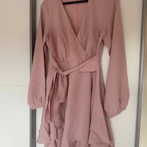 Ljusrosa omlottklänning med volang - Supersöt ljusrosa klänning med omlottliv, v-ringning och knytband i midjan. Aldrig använt då den var för stor