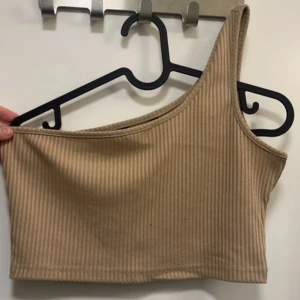 Beige ribbad one shoulder topp - Säljer en beige ribbad one shoulder topp. Toppen är croppad och har en tight passform med snygg ribbad struktur. Perfekt för att skapa en trendig och stilren look.