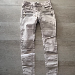 Beige skinny jeans med dragkedjor - Säljer ett par beige skinny jeans med fyra dekorativa dragkedjor framtill. Byxorna har en smal passform och normal midja, perfekta för en trendig och stilren look.
