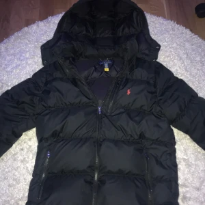 Svart pufferjacka från Polo Ralph Lauren - Säljer en svart pufferjacka från Polo Ralph Lauren med huva och dragkedja. Jackan har lång ärm och klassisk röd logga på bröstet. Perfekt för kyliga dagar och riktigt skön att bära. Jackan passar till längden 170 ungefär😀