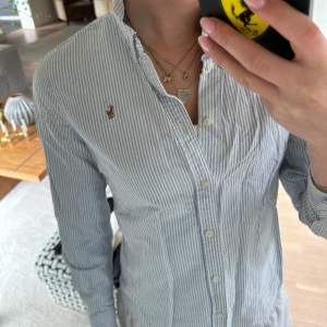 Blåvit randig skjorta från Ralph Lauren - Klassisk blåvit randig skjorta från Ralph Lauren med broderad logga på bröstet. Skjortan har lång ärm, knappar framtill.