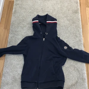 Mörkblå hoodiejacka från Moncler - Säljer en mörkblå hoodiejacka från Moncler med dragkedja och huva. hoodie har vita och röda ränder på huvan samt Moncler-logga på ärmen. Praktiska fickor framtill och ribbade muddar. Perfekt för dig som gillar stilren design och streetwear. det har en hål i vänstra sidan