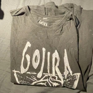 Svart Gojira t-shirt med tryck - Svart t-shirt med stort vitt Gojira-tryck på bröstet och motiv av en knytnäve. Klassisk rund hals och korta ärmar. Perfekt för dig som gillar bandmerch och vill ha en cool statement-tröja.