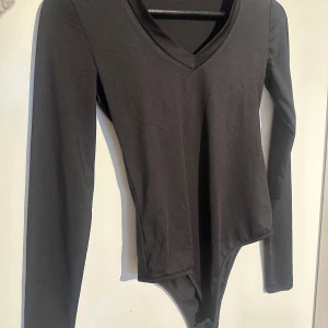 Svart långärmad bodysuit med v-ringning - En svart långärmad bodysuit med v-ringning. Tight passform och tillverkad i ett stretchigt material som sitter snyggt på kroppen. Perfekt att styla med jeans eller kjol.