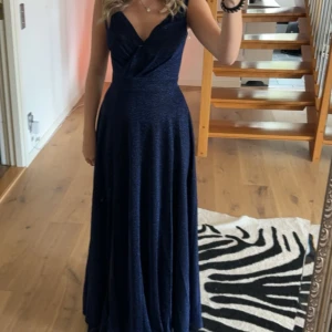 Mörkblå långklänning med slits - Säljer en elegant mörkblå långklänning med v-ringning och breda axelband. Klänningen har en markerad midja och en hög slits framtill som ger en snygg touch. Perfekt för bal eller fest! Materialet har en lätt glittrig finish. Endast använd 1 gång.