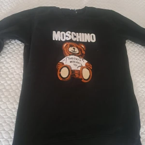  sweatshirt Moschino  - Svart sweatshirt från Moschino med stort vitt logotryck och en söt nalle på framsidan. Tröjan har rund hals och långa ärmar. Perfekt för dig som gillar statement-plagg och lekfull design. Passar M/L, nyskick 