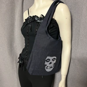 Mörkblå axelväska med Misfits-tryck - Säljer en mörkblå axelväska i jeansmaterial med Misfits-logga och ikoniskt dödskalletryck. Väskan har bred axelrem och en praktisk ficka framtill. Perfekt för dig som gillar alternativ stil och vill sticka ut. Helt ny.