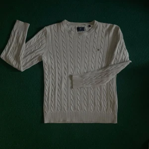 Vit kabelstickad tröja från GANT - Säljer en klassisk vit kabelstickad tröja från GANT i bomull. Tröjan har rund halsringning, långa ärmar och GANT-logga broderad på bröstet. Perfekt för lager-på-lager och passar till många olika stilar.