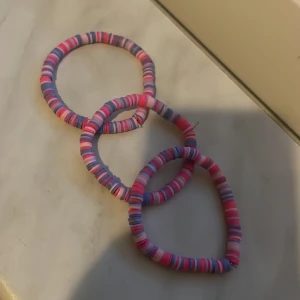 Färgglada armband i rosa och lila - Säljer tre elastiska armband i mjukt tyg med randigt mönster i rosa, lila och vitt. Perfekta som accessoar för att liva upp din outfit. Passar både till vardag och när du vill ha något extra färgglatt.