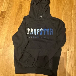 Svart hoodie från Trapstar - Säljer en svart hoodie från Trapstar med blå och vit logga på bröstet. Tröjan har huva med dragsko och en stor magficka. Perfekt för dig som gillar streetwear och vill ha något bekvämt och snyggt.