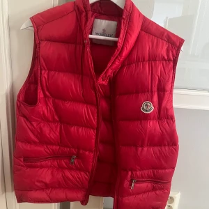 Röd dunväst från Moncler, storlek 2 motsvarar S. Pris kan diskuteras - Snygg röd dunväst från Moncler med dragkedja framtill och två fickor med dragkedja. Västen har Moncler-logga på bröstet och en quiltad design som ger en sportig look. Perfekt för lager-på-lager under kyliga dagar.
