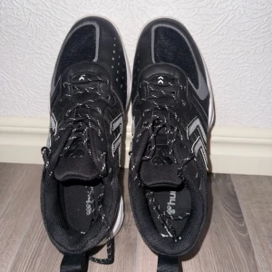 Svarta sneakers från Hummel - Säljer ett par svarta sneakers från Hummel med vita och grå detaljer samt vit sula. Skorna har snörning och är tillverkade i syntetmaterial med meshpaneler för extra andningsförmåga. Perfekta för träning eller vardagsbruk.