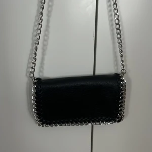 Svart axelväska med kedja från Tiamo - Snygg svart axelväska från Tiamo med silverfärgad kedja som axelrem och dekor runt kanterna. Väskan är rektangulär och har ett stilrent, modernt utseende. Perfekt för att lyfta din outfit med en cool detalj.