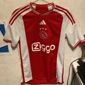 Ajax Amsterdam hemmatröja Ziggo - Ajax hemmatröja från Adidas med Ziggo som sponsor. Klassisk röd och vit design med klubbmärke och tre stjärnor på bröstet. Kortärmad modell med vita ränder på axlarna och diskreta ränder i tyget. Perfekt för fotbollsfans! Tröjan är äkta. 