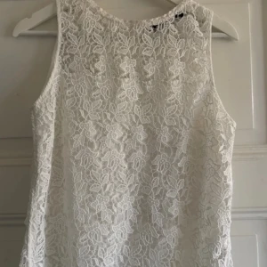 Vit spetslinne med blommönster - Säljer ett vitt ärmlöst linne med vackert blommigt spetsmönster. Perfekt för dig som vill ha en romantisk och somrig look. Linnet har rund halsringning och är helfodrat under spetsen.