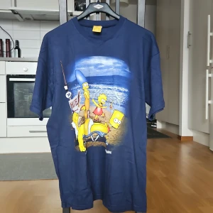 Blå The Simpsons t-shirt - Mörkblå t-shirt med stort tryck av The Simpsons på stranden. 