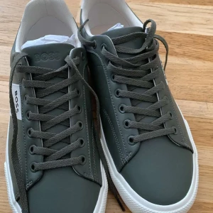 Gröna sneakers från BOSS med vita detaljer - Snygga gröna sneakers från BOSS med vita detaljer på häl och sidor. Skorna har rund tå, platt sula och klassisk snörning. Perfekta för dig som gillar stilrena och moderna sneakers med diskret logga på sidan och bak.