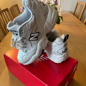 Vita New Balance 608 sneakers - Säljer ett par klassiska vita New Balance 608 sneakers med chunky sula och svarta detaljer. Skorna har snörning och är tillverkade i syntetmaterial med meshpaneler för extra komfort. Perfekta för en sportig och trendig look.