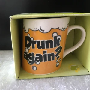Mugg med text 'Drunk again?' från Crazy Mugs - Rolig mugg i vitt och orange med texten 'Drunk again?' och bubblor som dekoration. Muggen levereras i originalförpackning med grönt mönster och illustrationer av olika muggar. Perfekt som present eller till samlingen.