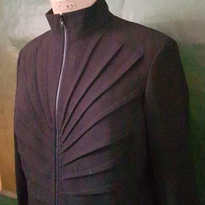 Svart jacka med veckad design och dragkedja - Säljer en svart jacka med unik veckad design framtill och hög krage. Jackan har lång ärm och stängs med dragkedja. Passformen är normal och materialet känns som ull eller liknande tyg. Perfekt för dig som gillar stilrena och annorlunda plagg.