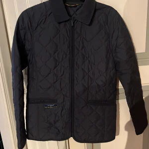 Mörkblå quiltad jacka från Henri Lloyd - Säljer en mörkblå quiltad jacka från Henri Lloyd med dragkedja framtill och klassisk krage. Jackan har två fickor med ribbade detaljer och diskret logga på framsidan. Perfekt för kyliga dagar.