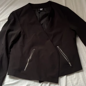 Svart kavaj med dragkedjor - Säljer en svart kavaj från H&M med två sneda dragkedjor framtill. Jackan har lång ärm och en stilren, minimalistisk design utan synliga knappar. Perfekt för dig som gillar en modern och enkel look.