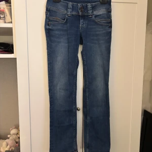 Blå straight jeans Venus low waist - Säljer ett par klassiska blå jeans från pepe jeans modell Venus med straight fit och låg midja. Jeansen har dubbla knappar i midjan, fem fickor och snygga detaljer på bakfickorna. Perfekta för en avslappnad och tidlös look.