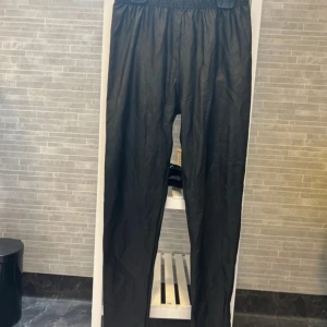 Svarta byxor i fuskskinn - Säljer ett par svarta byxor i fuskskinn med resår i midjan. Byxorna har en rak passform och är tillverkade i polyester och elastan, vilket gör dem både bekväma och snygga. Perfekta för dig som vill ha en trendig look.