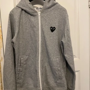 Grå hoodie från Comme des Garçons Play - Grå hoodie med dragkedja från Comme des Garçons Play. Tröjan har en svart hjärtdetalj med ögon på bröstet och en större hjärtdetalj på ryggen. Utrustad med huva och två fickor framtill. Liten fläck nertill och saknar snören, annars kanon skick!