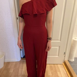 Röd one shoulder jumpsuit med volang - Oanvänd 34-38 Säljer en snygg röd jumpsuit med one shoulder och volangdetalj över axeln. Byxdressen har en tight passform upptill och vida ben. Perfekt för dig som vill sticka ut med en trendig look.