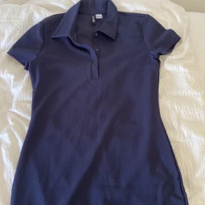 Marinblå pikéklänning från H&M - Säljer en enkel och stilren marinblå pikéklänning från H&M. Klänningen har korta ärmar, klassisk krage och knappar framtill. Perfekt för en avslappnad och sportig look.