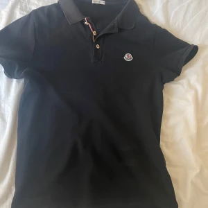 Svart piké t-shirt från Moncler - Säljer en svart piké t-shirt från Moncler med klassisk krage och knappslå med röd, vit och blå detalj. Moncler-logga broderad på bröstet. Perfekt för en stilren och sportig look. Storlek S/M
