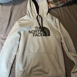 Ljusgrå hoodie från The North Face - Säljer en ljusgrå hoodie från The North Face med svart logga på bröstet. Tröjan har huva med svarta snören och en stor magficka framtill. Perfekt för dig som gillar enkel och stilren streetwear.