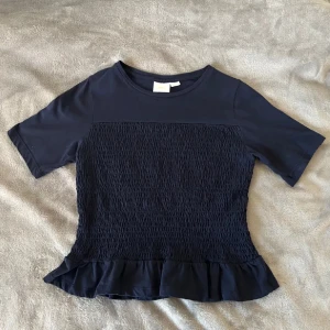 Mörkblå smockad topp - Säljer en mörkblå topp med korta ärmar. Toppen har smockad framsida och en volangkant nedtill som ger en söt touch. Perfekt till jeans eller kjol för en avslappnad stil.
