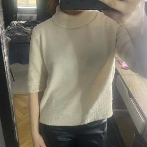 Vit stickad polotröja - Säljer en krämvit stickad polotröja med kortare ärmar och lös passform. Tröjan har en hög krage och är perfekt för lager-på-lager-stil. Enkel och stilren design som passar till mycket.