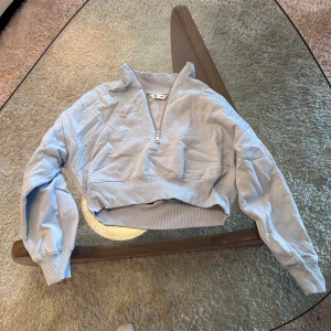 Ljusblå croppad sweatshirt med dragkedja - Säljer en ljusblå croppad sweatshirt med halv dragkedja och ribbade muddar. Tröjan har en hög krage och är perfekt för en avslappnad stil. Passar dig som gillar bekväma och trendiga plagg.