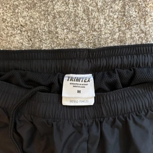 Svarta träningsshorts/badbyxor från Trimtex - Svarta träningsshorts från Trimtex med elastisk midja och snörning. Lätta och luftiga med diskret logga på vänster ben. Perfekta för löpning eller gym. Från Försvarsmakten 