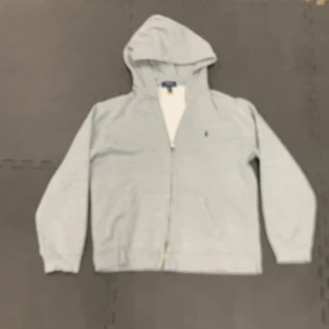 Grå hoodie från Polo Ralph Lauren XL - Snygg ljusgrå hoodie från Polo Ralph Lauren i storlek 170. Tröjan har dragkedja, huva och klassisk broderad logga på bröstet. Tillverkad i mjuk bomullsmix, perfekt för chill dagar. Långa ärmar och känguruficka framtill. | Denna tröja är helt autentisk! |