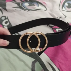 Svart bälte med gulddetaljer från SHEIN - Stilrent svart bälte från SHEIN med två runda guldfärgade ringar som spänne. Bältet är gjort i syntetmaterial och har en klassisk, enkel design som passar till många outfits. Perfekt accessoar för att lyfta din look. Finns 11st hål för att spänna bältet, köpte på plick men visste inte att den var från shein +var den för lång på mig. Den är ca 98cm lång