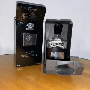 Creed Aventus 100ml Eau de Parfum - Lyxig parfym från Creed, Aventus, i en elegant 100ml glasflaska med svart och silverdetaljer. Flaskan har en stilren svart kork och levereras i en svart kartong med silverlogga och text. En ikonisk doft med exklusiv känsla.