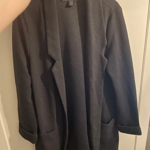 Svart kappa från Amisu, strl 36 - Svart lång kappa från Amisu i storlek 36. Jackan har en stilren design med slag, uppvikta ärmslut och två fickor framtill. Perfekt för dig som gillar en clean och enkel look. Materialet är mjukt och har en lätt ribbad struktur.